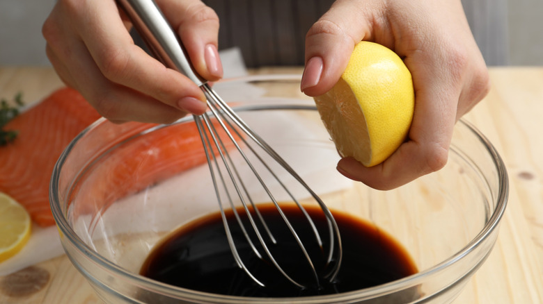 Hands whisking lemon juice into soy sauce in a glass bowl on a countertop with salmon in the background