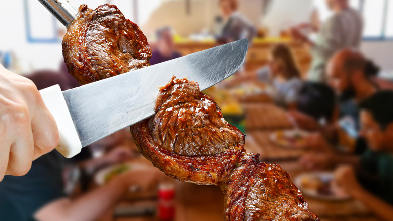 A knife slices picanha from a skewer, people out of focus in the background