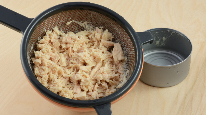 Empty tuna fish can and tuna in a strainer on a light wood countertop